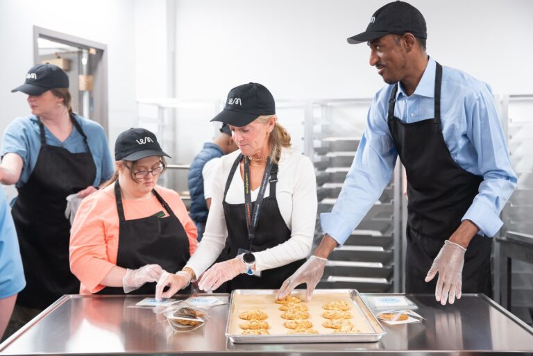 Four bakers gather around the prep table to decorate a tray of cookies