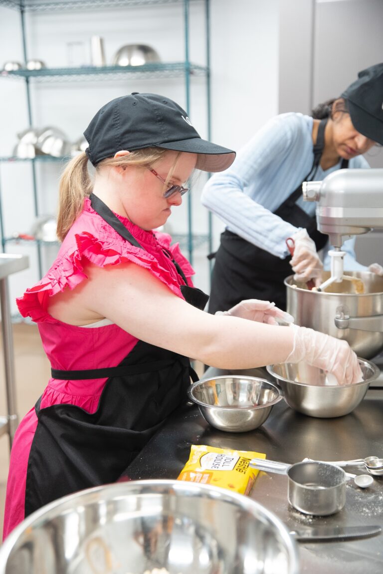 A baker in a pink ruffle top scooping ingredients at a bakery counter