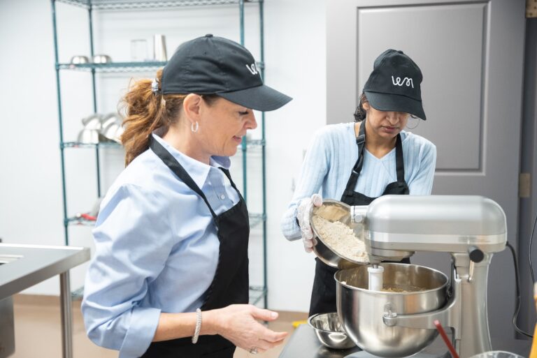 A staff member pouring flour into a stand mixer bowl as a baker watches