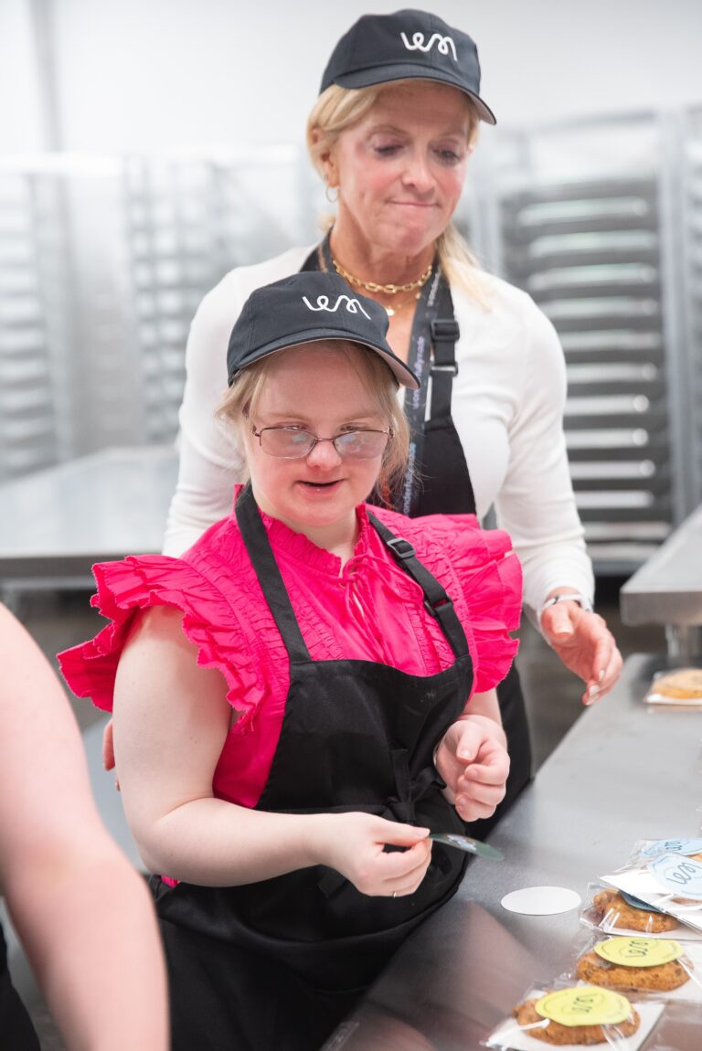 A young woman in glasses applies a label to a packaged cookie as a staff member watches